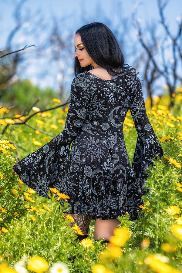 A woman with long dark hair stands in a field of yellow flowers, wearing the Mage Mini Dress by Melodia Designs, which features a black celestial print and wide bell sleeves. She looks over her shoulder amid bright greenery under a blue sky.