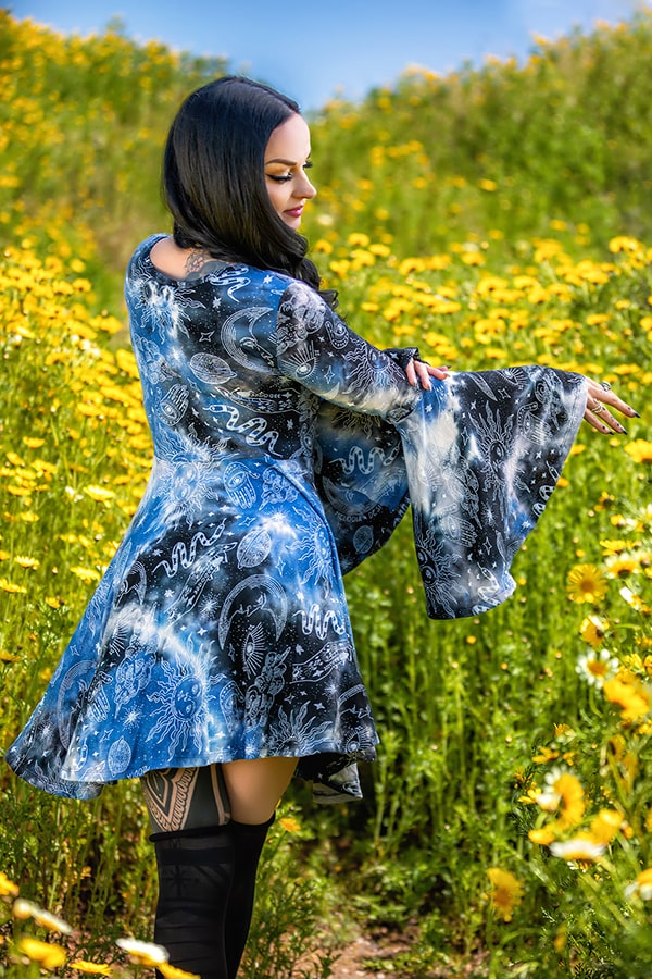A woman with long dark hair wears the Melodia Designs Mage Mini Dress, featuring blue and black bell sleeves and a flattering fit, as she stands in a bright field of yellow wildflowers, looking down with her arm extended.