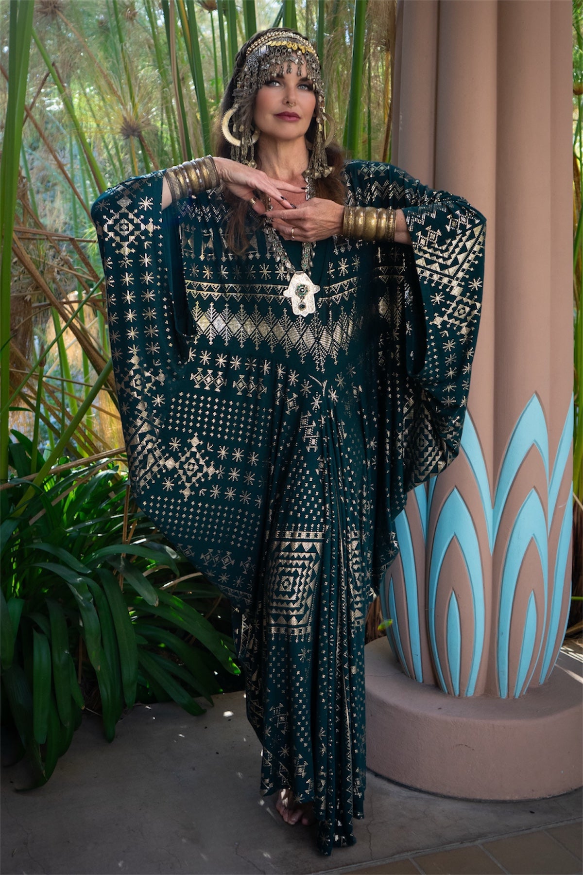 A person poses before tall plants and a blue-patterned column, wearing the Melodia Designs Alkimiya Contessa Caftan Malachite—a dark, patterned caftan with gold accents, deep V neckline, and batwing sleeves—styled with bold jewelry and an ornate headpiece.