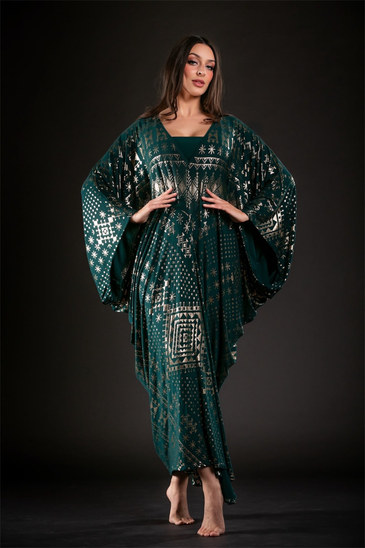 A woman poses barefoot against a dark background, wearing the Alkimiya Contessa Caftan Malachite by Melodia Designs—an elegant batwing-sleeve kaftan in dark green and gold with wide geometric patterns and a deep V neckline.