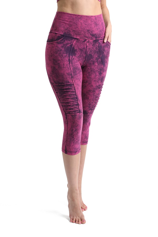 A person wears Melodia Designs Moto Capri 2.0 Grunge Tourmaline, high-waisted pink and purple tie-dye capri leggings with moto detailing, standing barefoot against a white background.