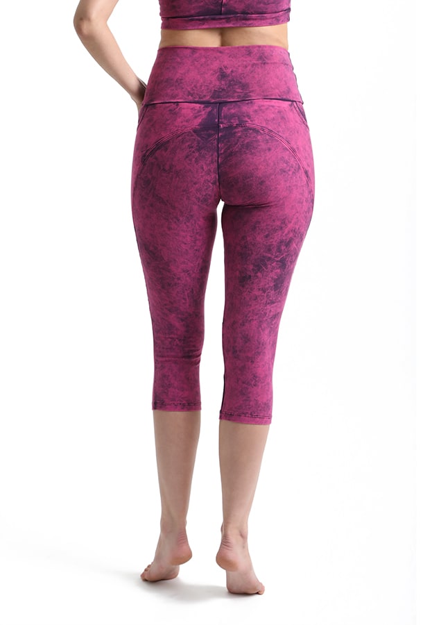 A person standing barefoot, seen from the back, wears Melodia Designs Moto Capri 2.0 Grunge Tourmaline leggings and matching sports top in a pink and purple marbled print against a white background.