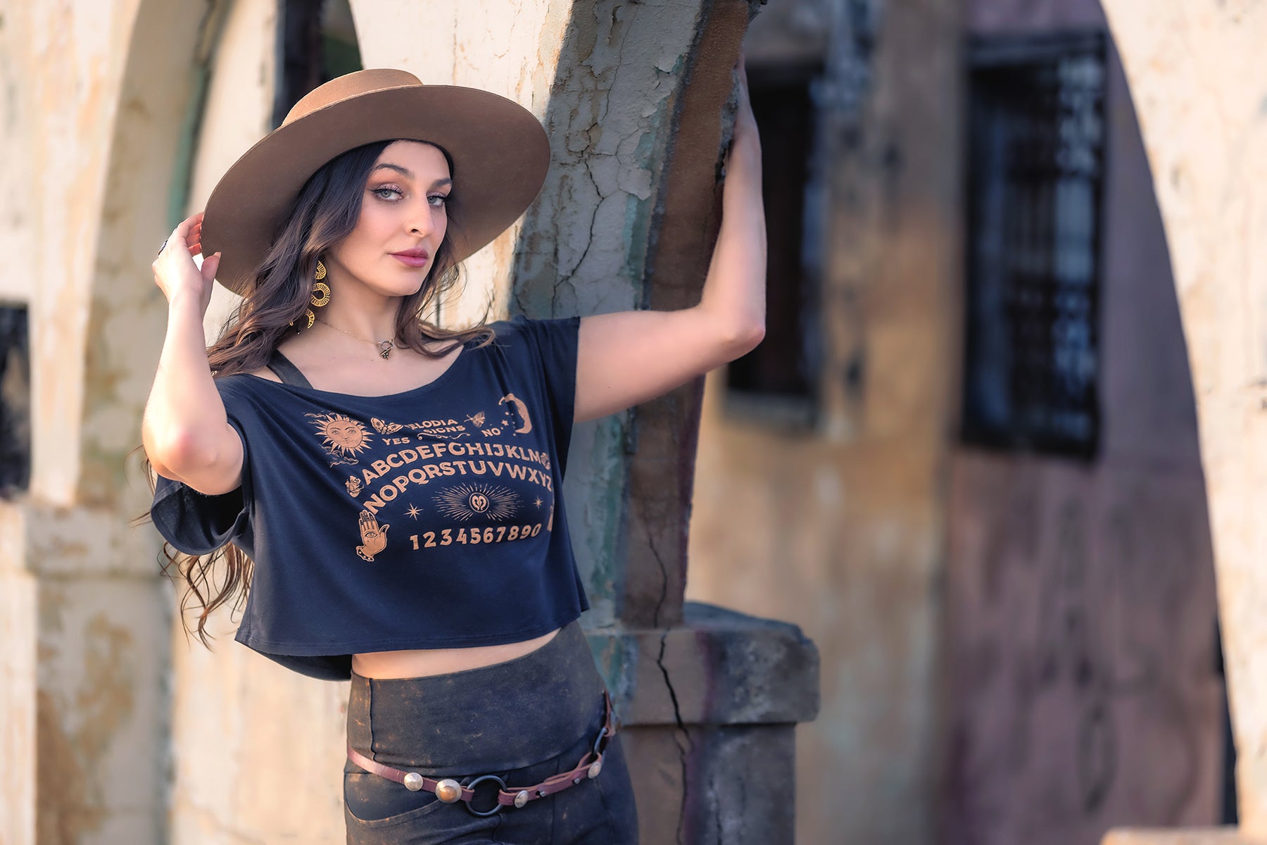 A woman wearing the Ouija Dede Crop by Melodia Designs and a wide-brimmed hat stands outdoors, leaning on a stone archway and looking confidently at the camera.