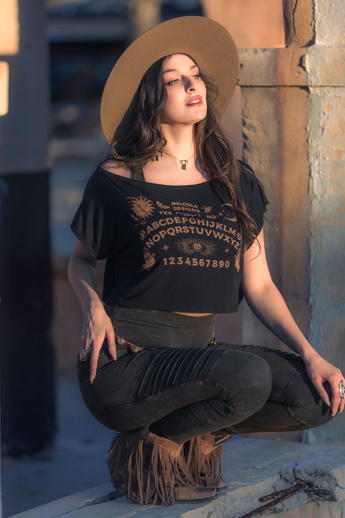 A woman in a wide-brimmed hat and the Melodia Designs Ouija Dede Crop with a mystical design sits outdoors on a fringed stool, gazing upward as sunlight highlights her face and hair.