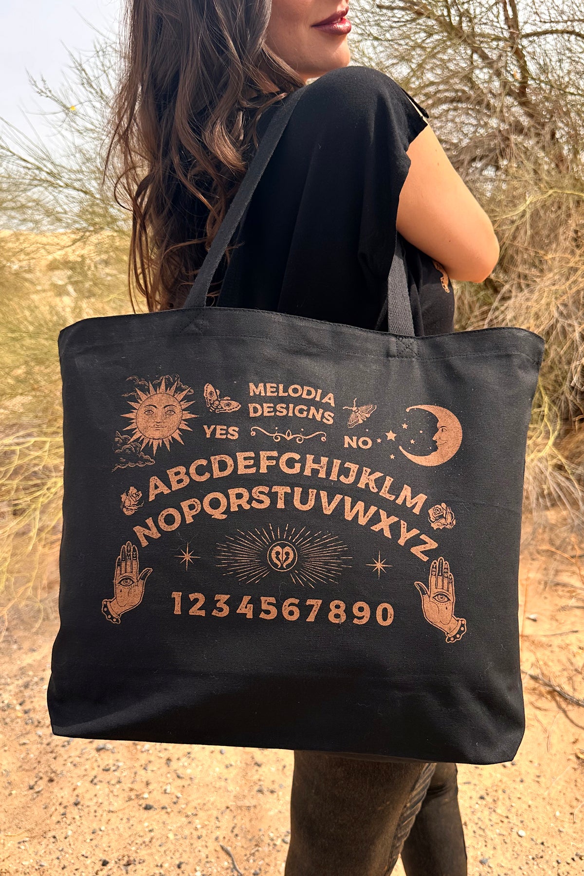 A woman outdoors holds the Ouija Tote Bag by Melodia Designs, featuring tan Ouija-inspired designs with alphabet, numbers, sun, moon, stars, two hands, and the words YES and NO.