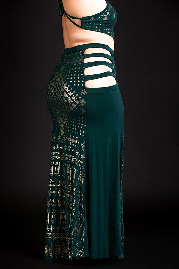 A person wearing the Melodia Designs Alkimiya Persephone Skirt Malachite, featuring eco-luxe fabric, dark green and gold geometric patterns, side cut-outs, and horizontal straps, stands in profile against a black background.