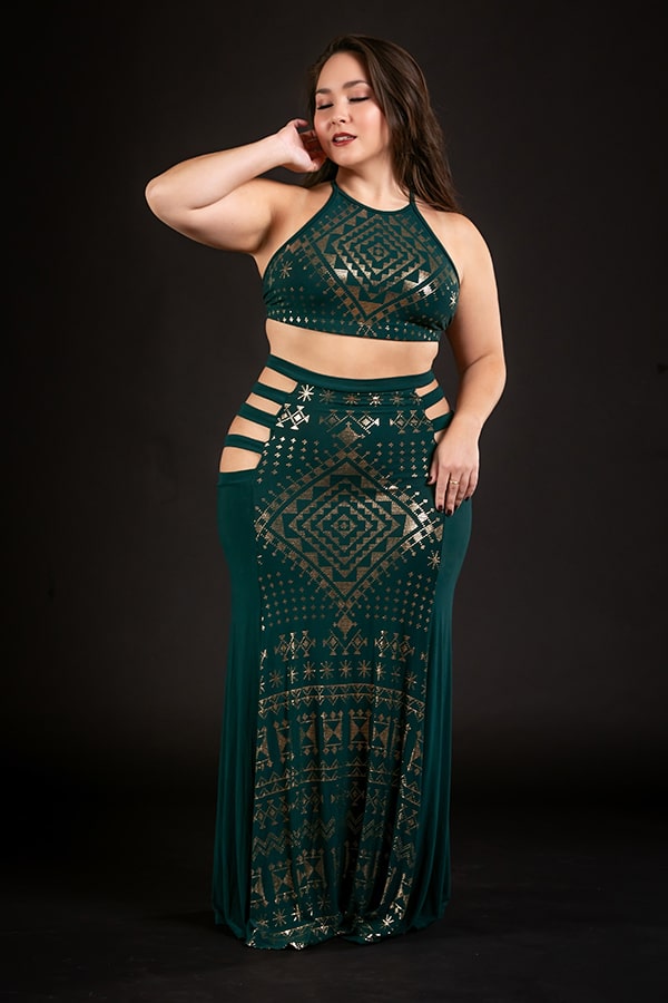 A woman poses confidently in the Melodia Designs Alkimiya Persephone Skirt Malachite—a dark green, sleeveless two-piece with gold geometric print and waist cut-outs—against a plain black background.