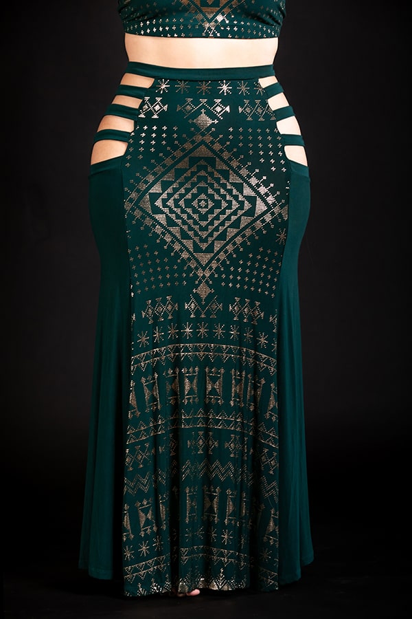 A person wears the Melodia Designs Alkimiya Persephone Skirt Malachite, a long eco-luxe dark green skirt with gold geometric and tribal Al-kimiya patterns, cut-out waist straps, and a matching cropped top, set against a black background.