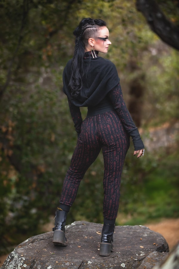 A person with long dark hair and black sunglasses stands on a rock outdoors, wearing the Melodia Designs Freydis Hoodie Runes over a patterned jumpsuit, paired with black gloves and platform boots. Trees and greenery are in the background.