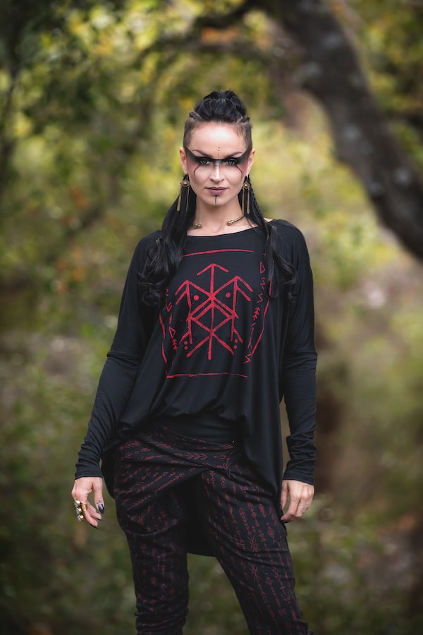 A person with long dark hair, dramatic makeup, and piercings stands outdoors in a black Melodia Designs Luna Long Sleeve Runes shirt featuring a red bindrune-inspired design, surrounded by blurred greenery.