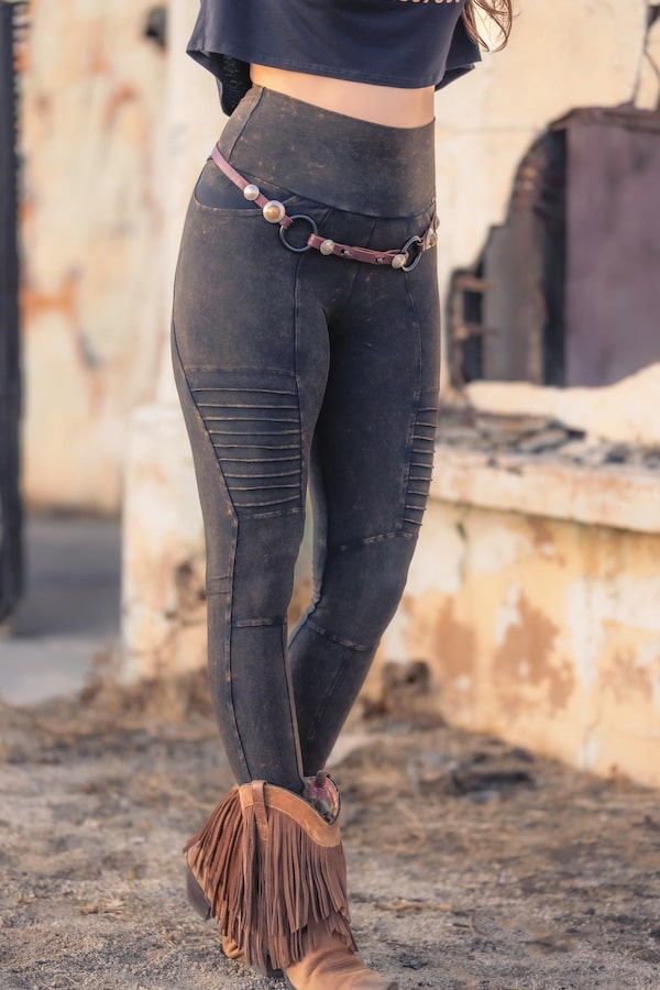 A person in Melodia Designs Rustique Moto Xtreme Pocket Legging, paired with a brown belt and tan fringed boots, stands outdoors before a weathered light wall.