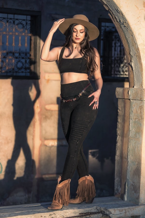 A woman in Melodia Designs Rustique Serena Bra 2.0 stands confidently under an archway, sunlight casting her shadow on the wall as she pairs the crop top with black leggings, a wide-brimmed hat, and fringe boots.