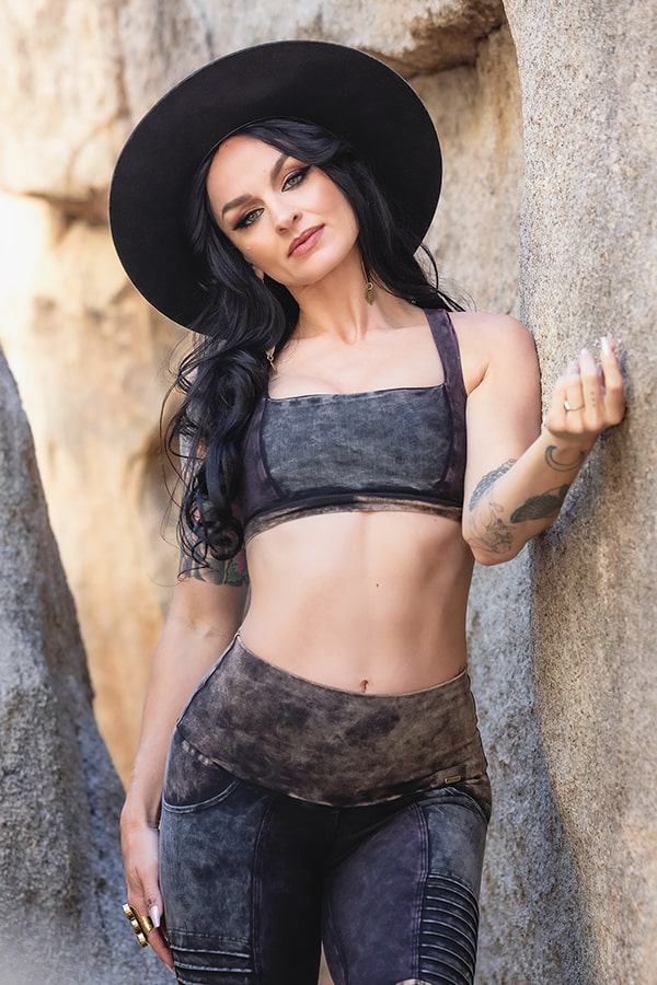 A woman with long dark hair smiles outdoors between large rocks, resting one hand on a rock while wearing the Serena Bra 2.0 Grunge by Melodia Designs—a dark, eco-luxe athletic top paired with a wide-brimmed black hat.