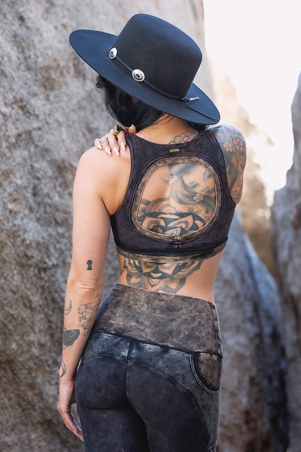 A person with long dark hair and tattoos stands outdoors between large rocks, wearing Melodia Designs’ Serena Bra 2.0 Grunge—a wide-brimmed black hat tops off their eco-luxe dark workout look.