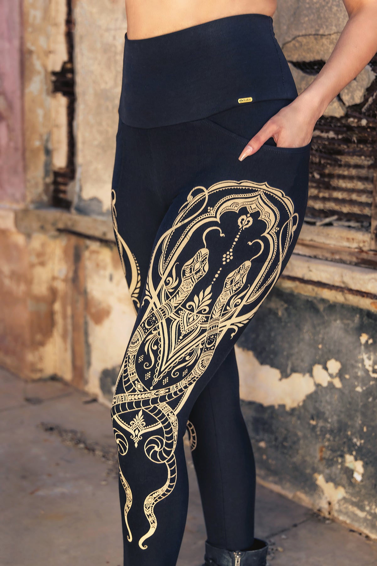 A person stands outdoors with one hand on their hip, modeling Melodia Designs Elsa Legging Serpent—high-waisted black leggings featuring intricate gold serpent designs. The face is hidden, and a worn, textured wall forms the background.