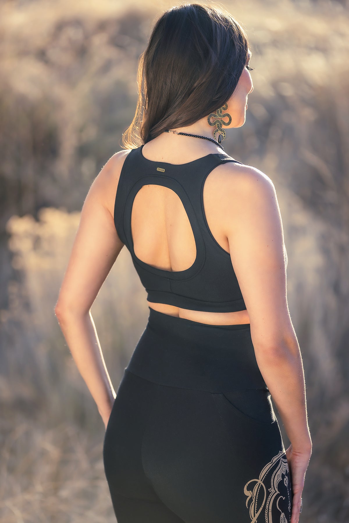 A woman with long brown hair, earrings, and a necklace stands outdoors in a black racerback sports bra and Melodia Designs Elsa Legging Serpent with a high-rise waistband. The background features blurred natural scenery.