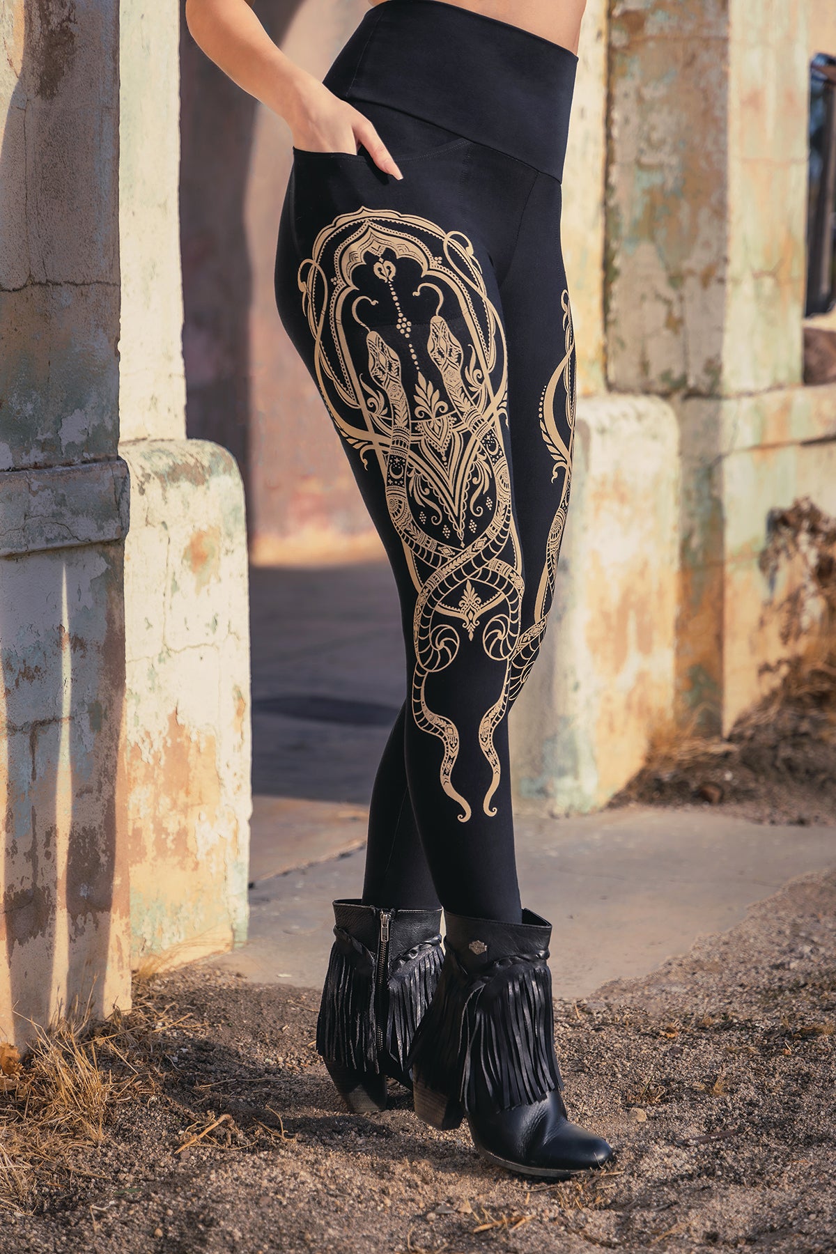 Wearing Melodia Designs Elsa Legging Serpent—detailed with a gold serpent motif and high-rise waist—paired with black fringe ankle boots, someone stands by a weathered outdoor wall, showing only their lower half.