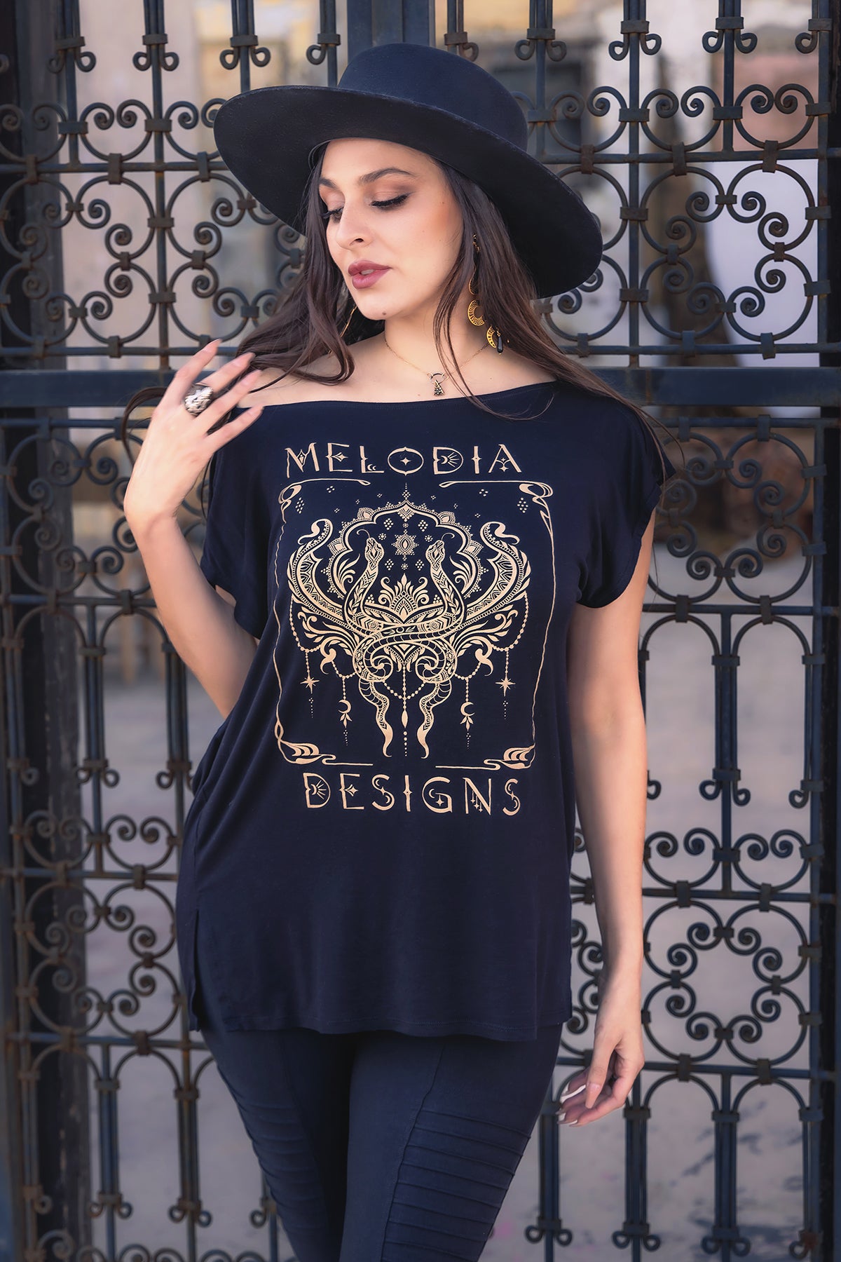 A woman in a wide-brimmed black hat and a black Melodia Designs Nouveau Serpent Tunic with a gold ornate graphic stands before a decorative iron gate, looking to the side with one hand on her hat. The tunic is crafted from eco-luxe fabric for comfort.