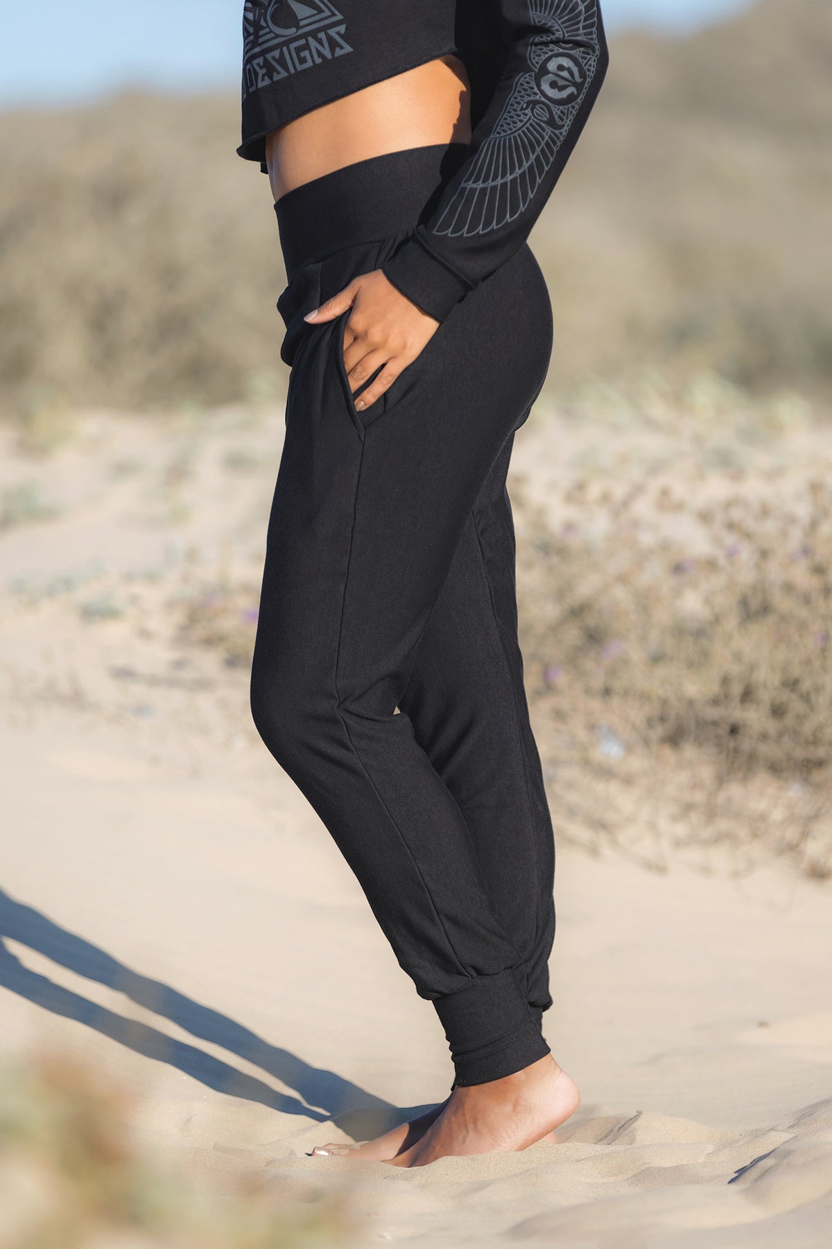A person stands barefoot on sand, wearing Melodia Designs' Shiva Wrap Jogger Black pants and a long-sleeve black top with patterned sleeves. The blurred background of dry grass and shrubs suggests eco-friendly comfort.