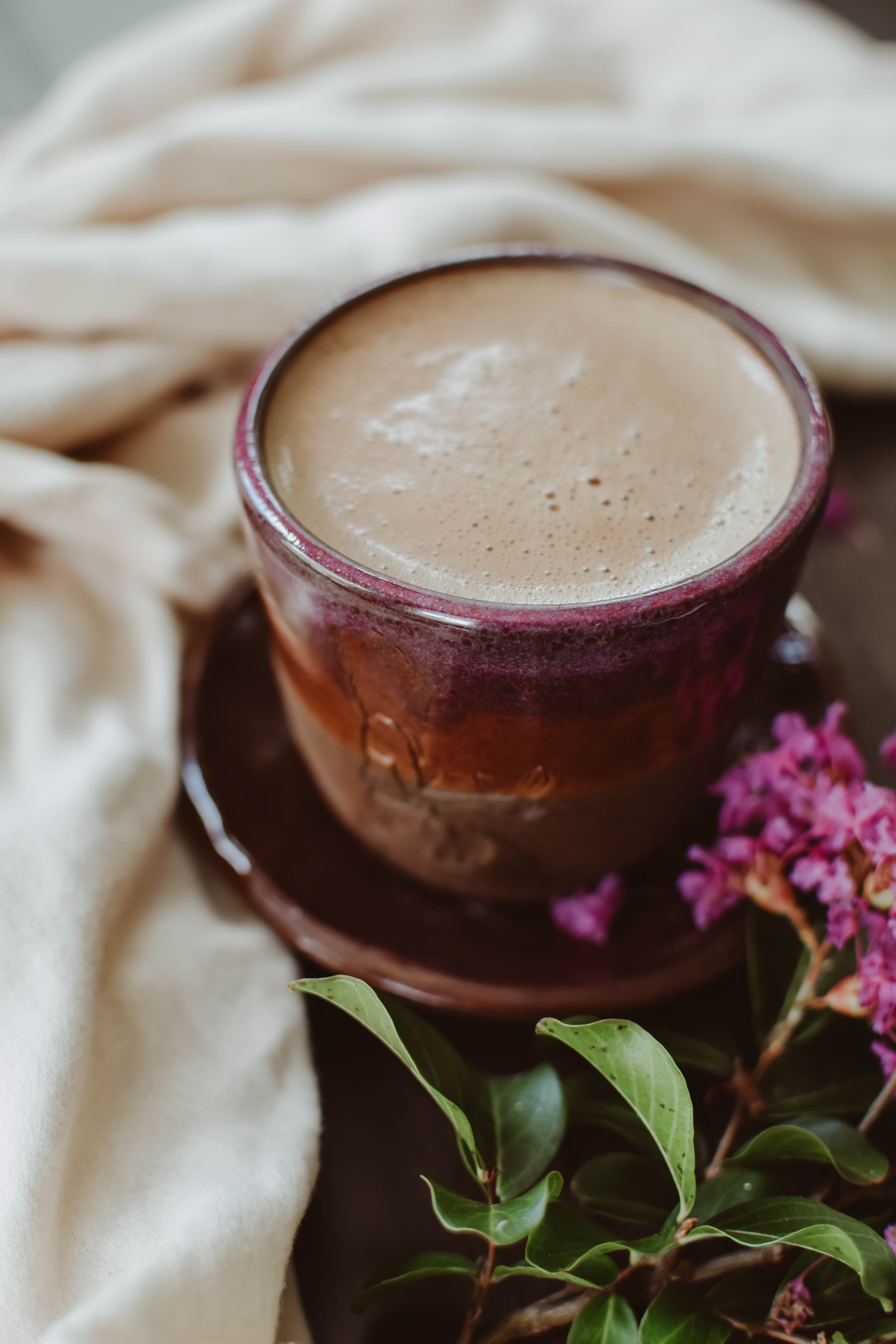 A Heartswell Sipping Chocolate mug by chocolita.com rests on a matching saucer, surrounded by a white cloth and pink flowers with green leaves.