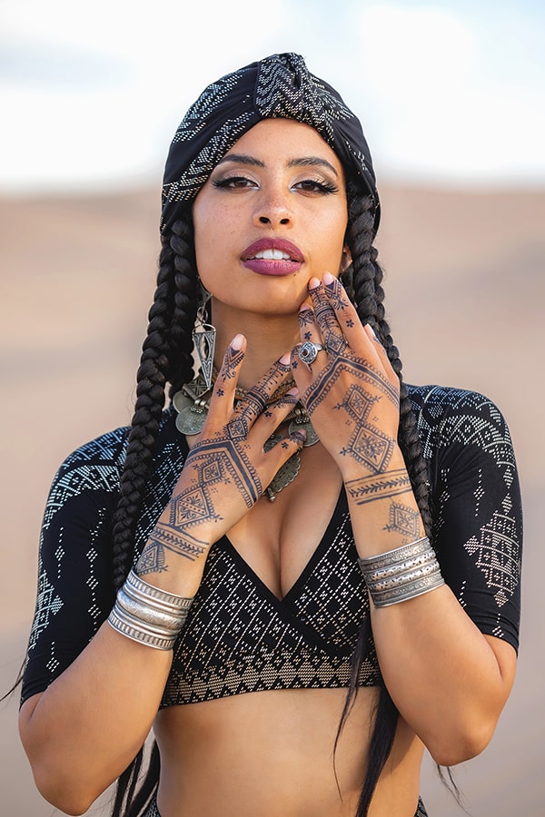 A woman with long braids poses outdoors in an Urban Turban by Melodia Designs. She wears a black and white patterned top, silver bracelets, large earrings, and intricate henna on her hands.