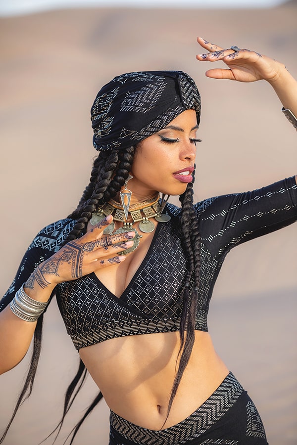 A woman with long braids and henna designs poses outdoors in a Melodia Designs Urban Turban, black patterned crop top, and statement jewelry—necklaces, bracelets, rings, and large earrings.