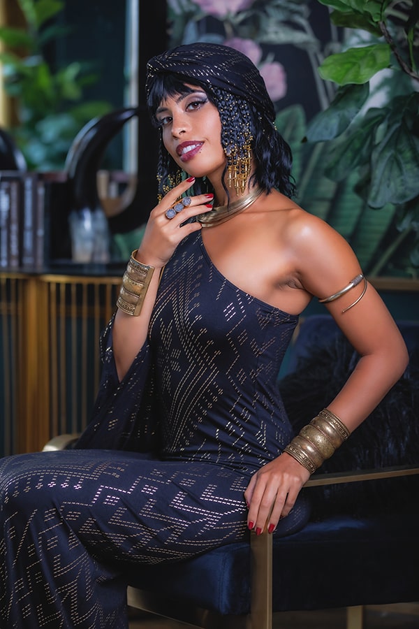 A woman in a black, one-shoulder patterned dress sits on a chair, wearing gold jewelry and the Melodia Designs Urban Turban Luxor. With short, face-framing twisted hair and bold makeup, she poses amid lush plants, hand gracefully touching her chin.