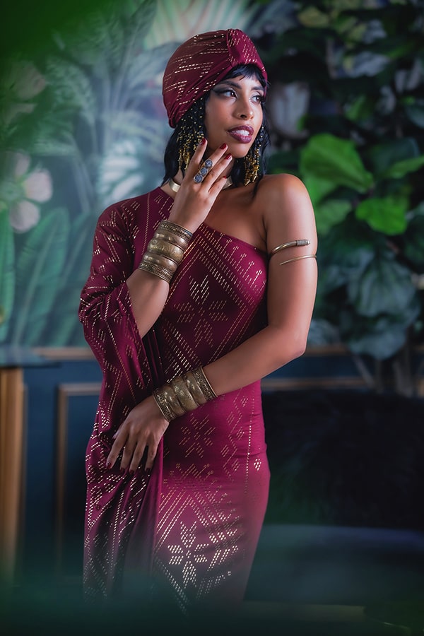 A woman in a maroon patterned dress and gold statement jewelry poses gracefully indoors, wearing the versatile Melodia Designs Urban Turban Luxor, styled elegantly against a backdrop of lush green plants.