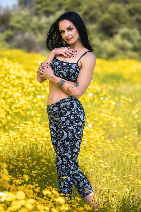 A woman with long dark hair smiles in a field of yellow wildflowers, wearing Melodia Designs Zoe Capri Alchemy yoga pants featuring white celestial patterns and deep pockets, paired with a crop top. Tattoos accent her arm against a blurred tree backdrop.