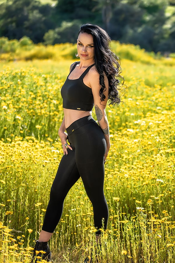 A woman wearing Melodia Designs’ Zoe Capri stands in a field of yellow wildflowers, posing with one hand on her hip as sunlight filters through green foliage in the background.