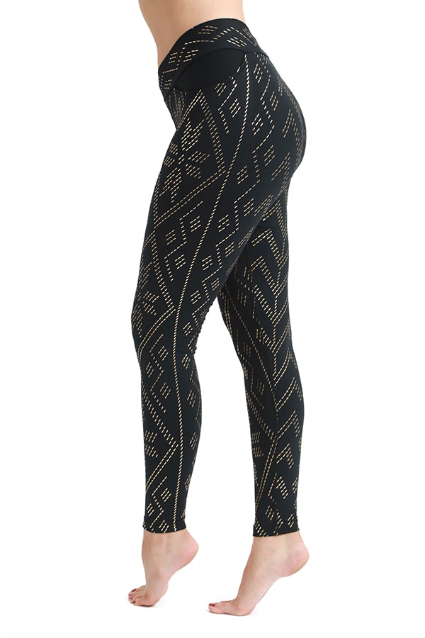A person poses barefoot in the Zoe Luxor Pocket Legging by Melodia Designs—high-waisted, full-length black leggings with a gold geometric design, crafted from high-performance stretch fabric, shown in side profile against a white background.