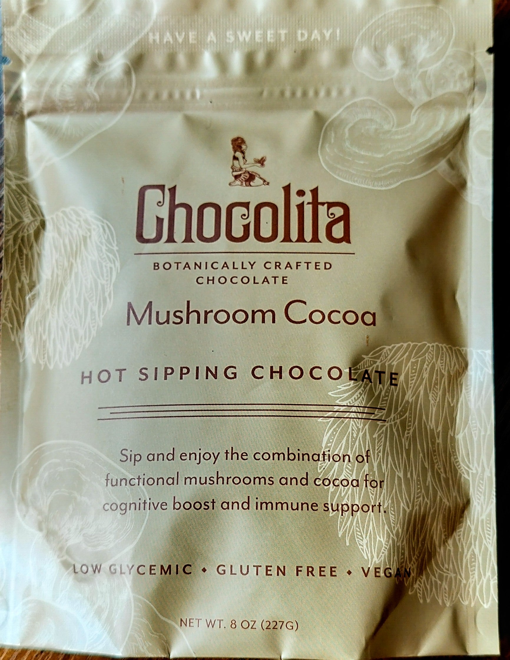 A beige packet of chocolita.com Mushroom Cocoa Sipping Chocolate features white illustrations and text highlighting botanical ingredients, medicinal mushrooms for cognitive and immune support, plus low glycemic, gluten free, and vegan benefits.