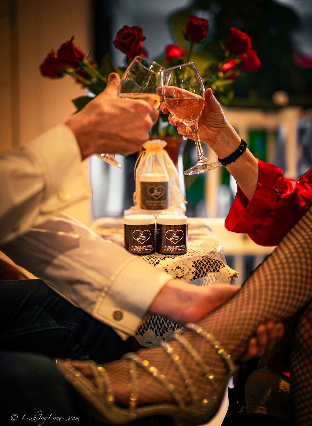 Two people share an intimate, romantic moment over a table with roses, a gift bag, chocolates, and Benoit Boutique's Love Body Butter—multi-purpose, all natural & edible in a 4oz jar. One wears a red blouse; the other wears white.