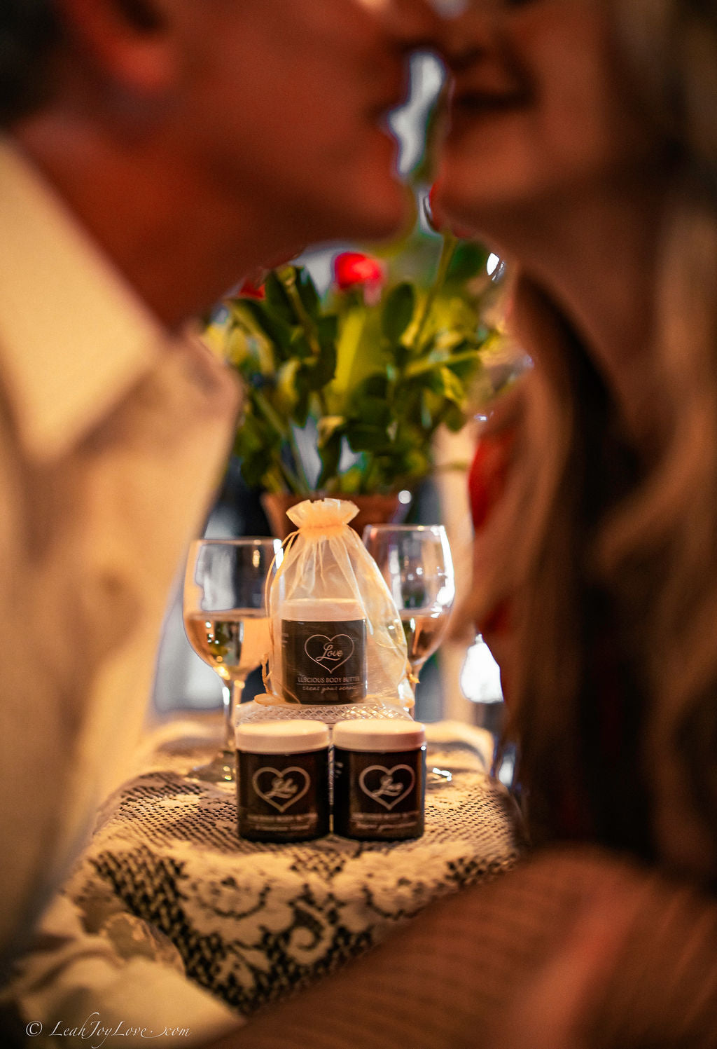 A couple leans in for a kiss, blurred in the foreground. In focus: two glasses of white wine, a Benoit Boutique Love Body Butter—Multi-Purpose, All Natural & Edible, 4oz jar with heart label—and a bouquet of flowers behind.