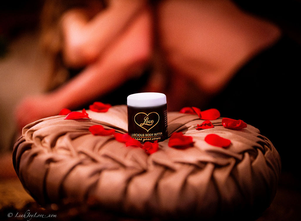 A 4oz jar of Benoit Boutique Love Body Butter—multi-purpose, all natural, and edible—rests on a textured cushion with red rose petals, while two shirtless people embrace in the soft-focus background for a sensual massage ambiance.