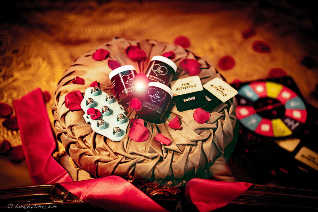 A gold cushion with rose petals, two Benoit Boutique Love Body Butter jars, a blue bell bracelet, FUN ACTIVITIES cards, chocolates, and a colorful game wheel are displayed on a golden surface with red ribbons.