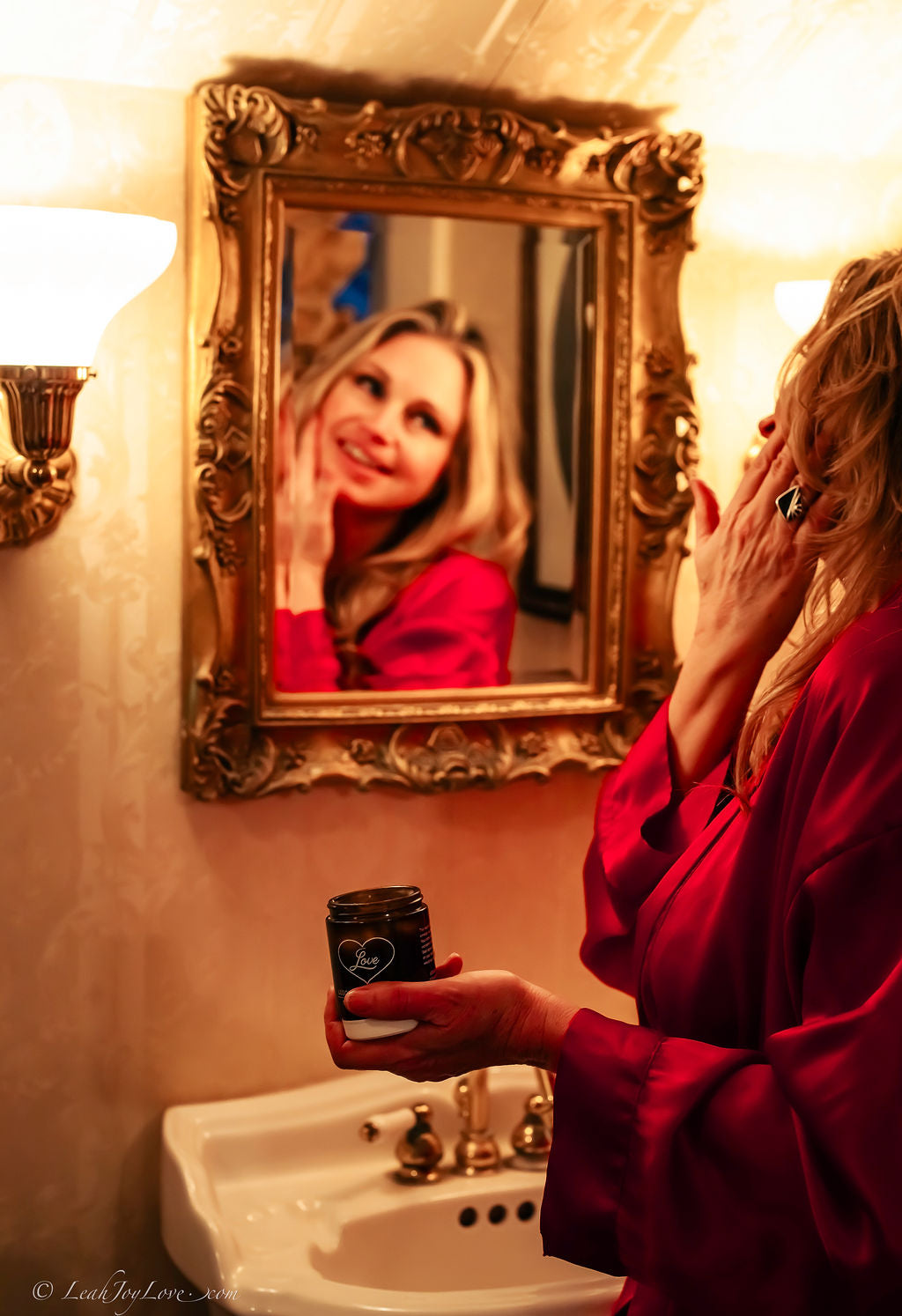 A woman in a red robe applies Benoit Boutique’s Love Body Butter—Multi-Purpose, All Natural & Edible (4oz)—to her face at the sink, gazing into a gold-framed mirror amid warm lighting and cozy, elegant bathroom decor.