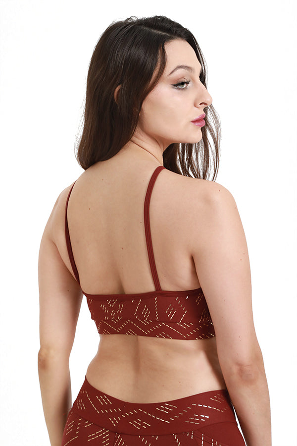 A woman with long brown hair models the Melodia Designs Kora Bra Luxor—a maroon sports bra with a geometric cutout pattern—standing backward and glancing over her shoulder against a white background.