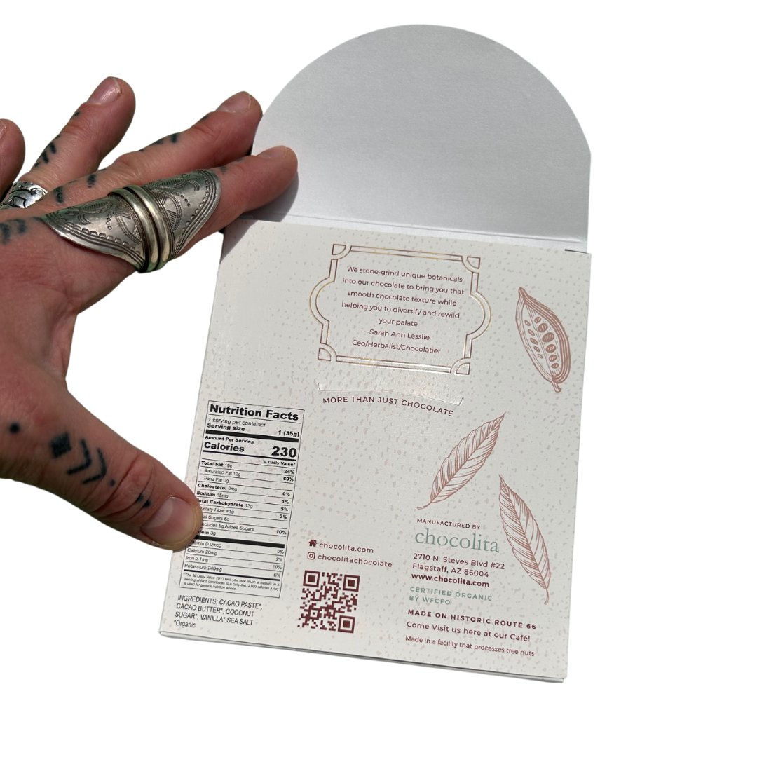 A tattooed hand with silver rings holds an open ECUADOR- Dark & Sweet bar from chocolita.com, revealing nutrition facts, contact info, and a QR code. The cream package is decorated with leaf illustrations.