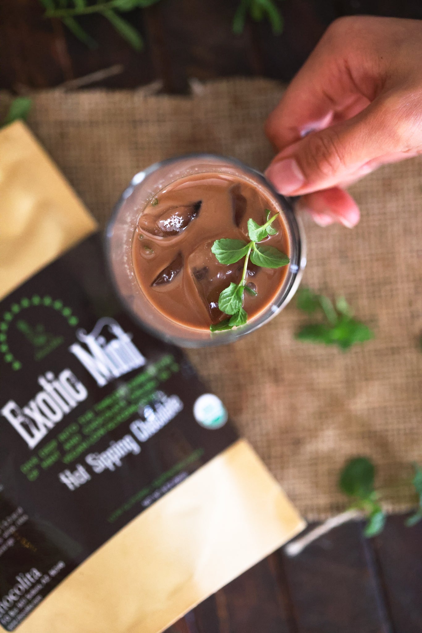 A hand holds a glass of iced vegan Exotic Mint Sipping Chocolate by chocolita.com, garnished with mint leaves. Below, a brown and beige chocolita.com packet is surrounded by scattered mint leaves on burlap.
