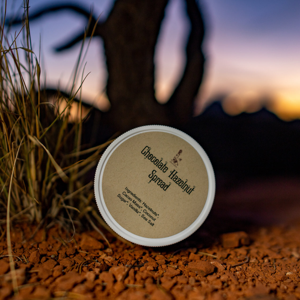 A jar of chocolita.com Hazelnut Chocolate Spread sits on reddish soil near dry grass, with a silhouetted tree and a colorful sunset sky in the blurred background.