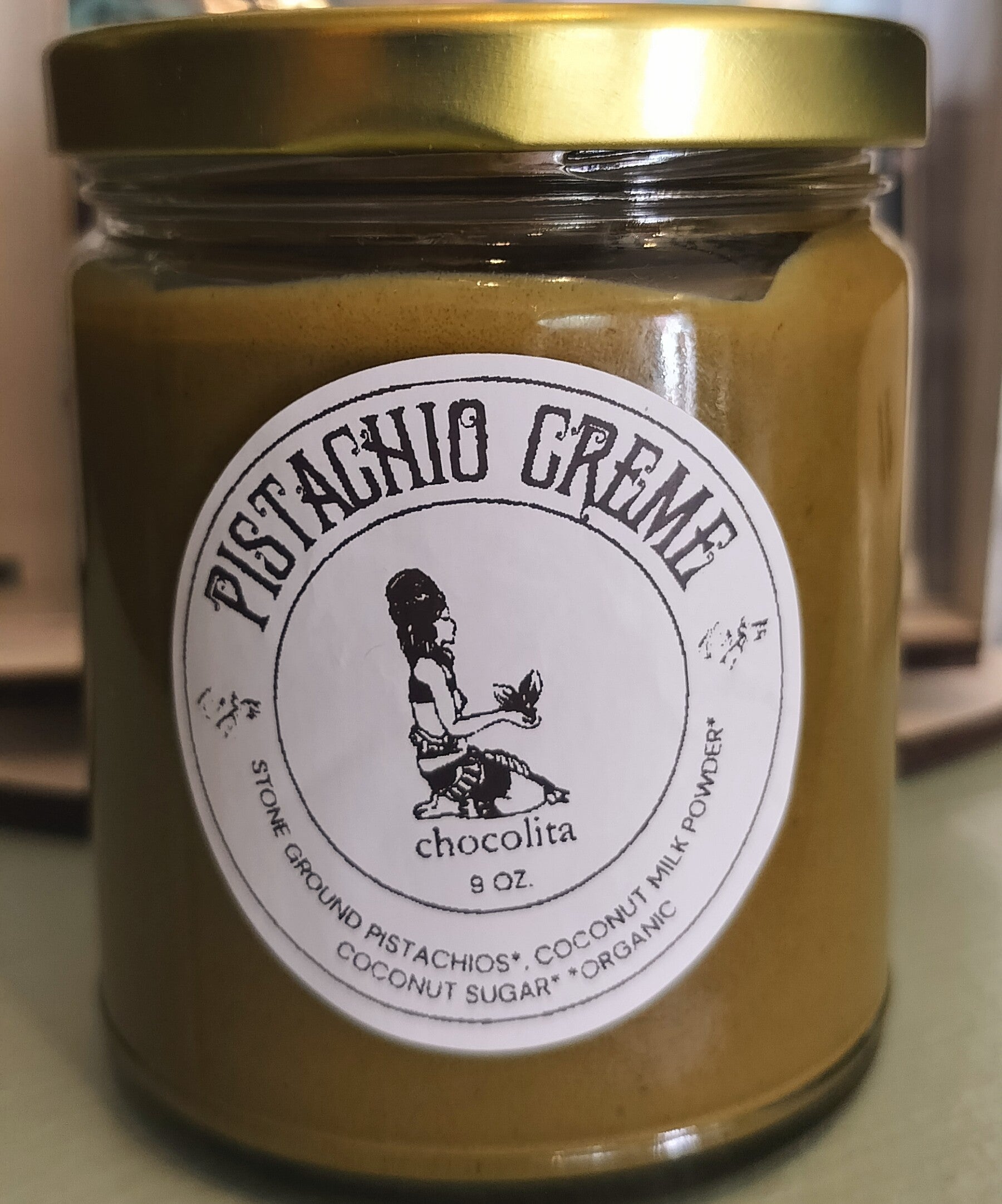 A 9 oz. jar of Pistachio Creme by chocolita.com, with a gold lid and a black-and-white label illustration of a seated person; contains stone ground pistachios, coconut mylk powder, coconut sugar, and organic ingredients.