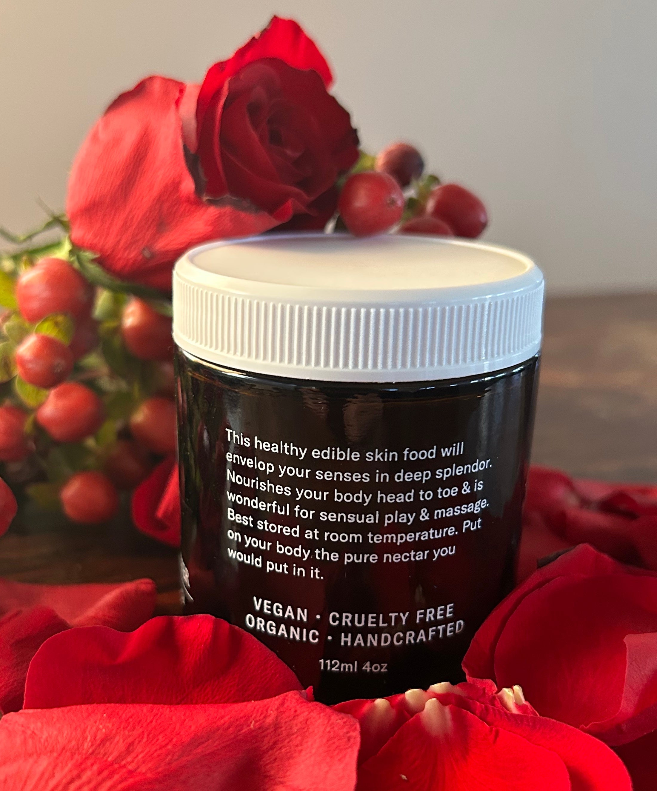 A 4oz jar of Benoit Boutique’s Love Body Butter—multi-purpose, all natural, and edible—rests among red rose petals and rose hips. The label highlights its nourishing benefits for skin care and sensual massages.