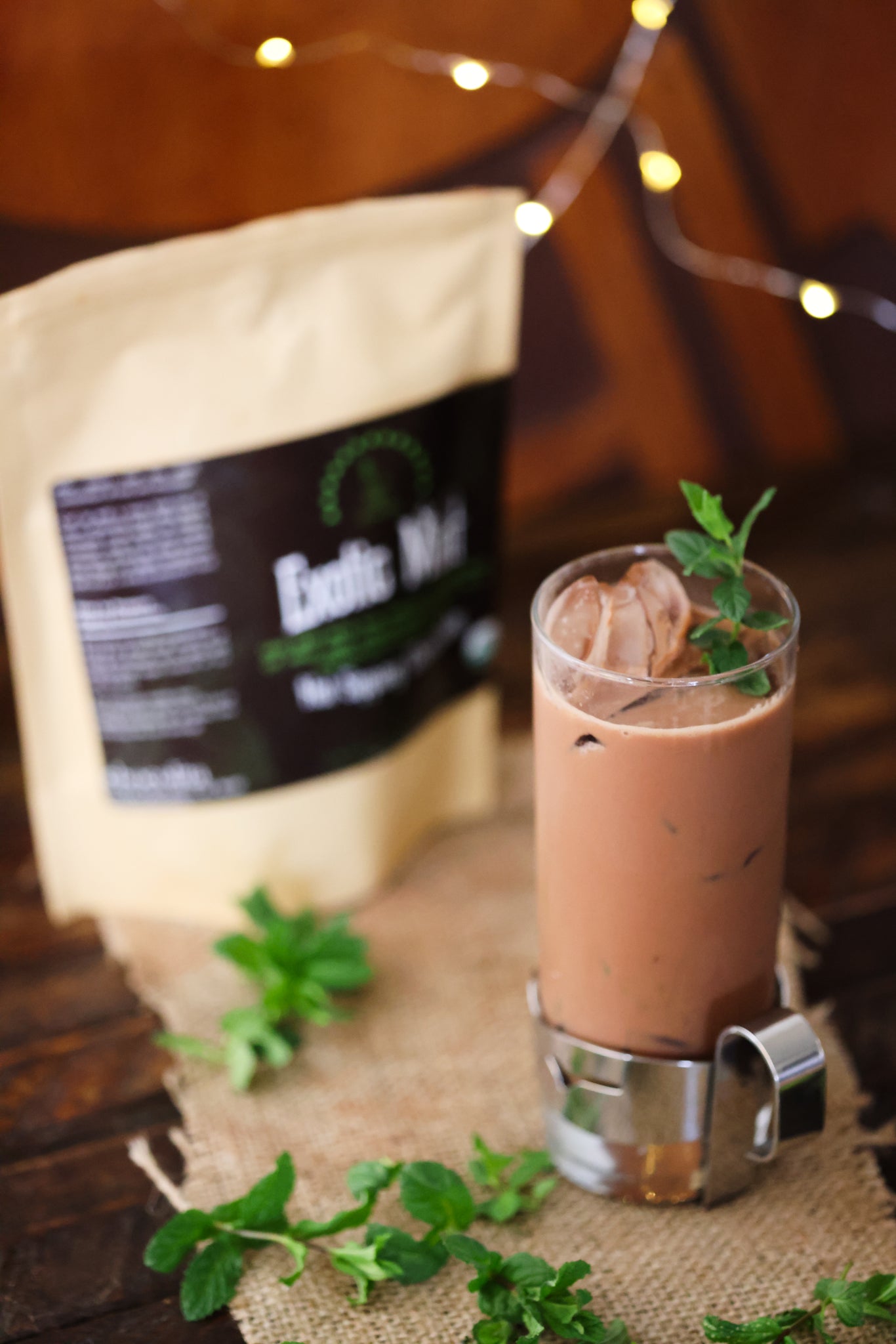 A tall glass of iced Exotic Mint Sipping Chocolate by chocolita.com, topped with mint and served beside its beige package and fresh leaves, rests on burlap as warm fairy lights glow, highlighting this low-glycemic treat.