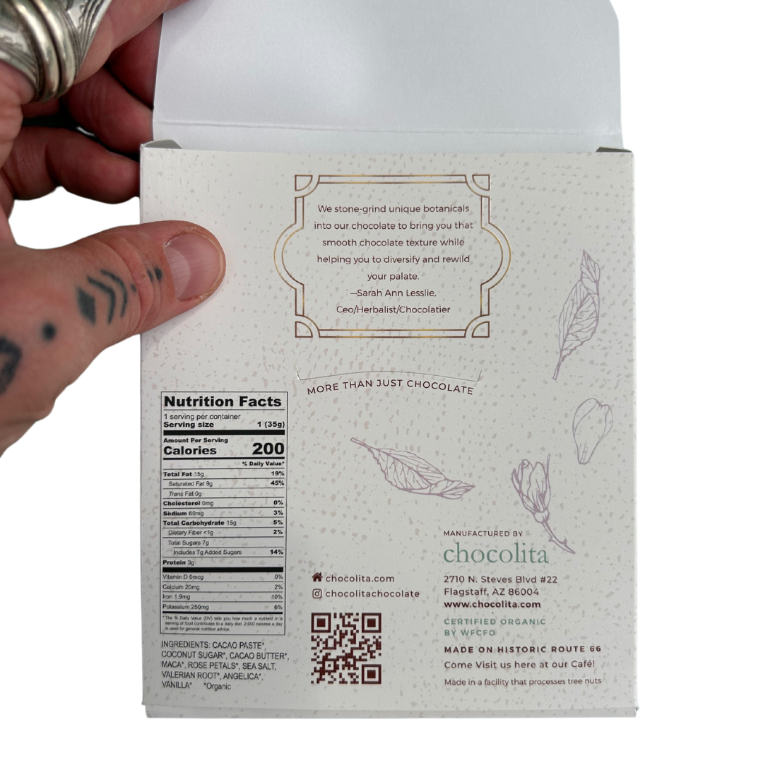 A tattooed, ringed hand holds the back of an EASE- Moontime Rose bar by chocolita.com, showing nutritional details, brand info, a quote, and illustrated cacao leaves with Female Cycle Herbs.