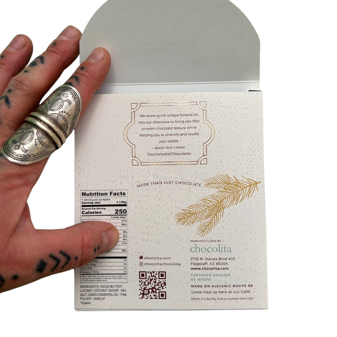 A tattooed hand with a silver ring holds an open POWER- Pine Pollen & Lemon raw white chocolate box. The back displays nutrition facts, a quote, chocolita.com branding, and a gold pine pollen branch illustration.