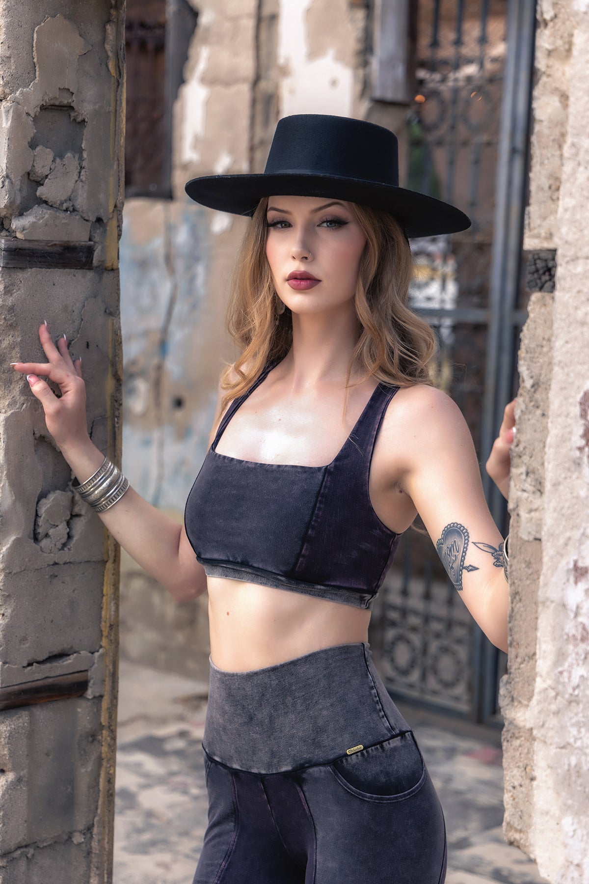 Wearing the River Rock V2 Serena Bra by Melodia Designs, a woman stands beside a weathered stone wall, her bird tattoo visible on her arm. She completes the look with a black wide-brim hat and matching pants for an eco-luxe style.