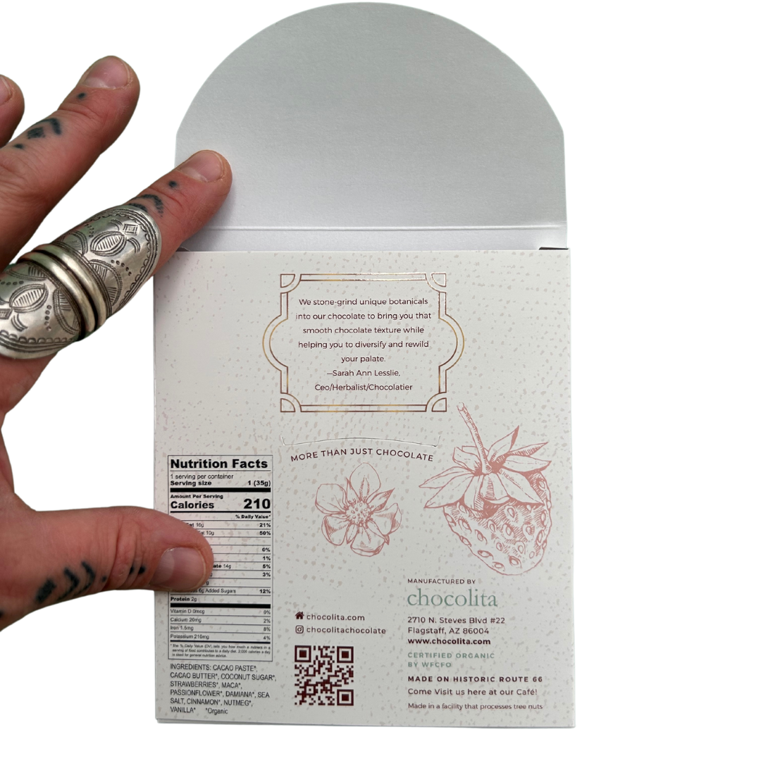 A hand with rings holds an open APHRODISIA- Strawberry Passion by chocolita.com, revealing nutrition and ingredient details with maca and aphrodesia herbs, botanical art, a QR code, and a handcrafted chocolate message on the inner white chocolate flap.