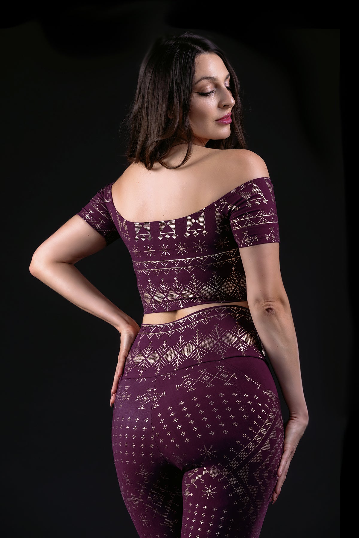 A woman poses against a black background in the Melodia Designs Alkimiya Allie Crop Top Aubergine-Gold, featuring eco-luxe fabric with beige geometric patterns—perfect for stylish layering.
