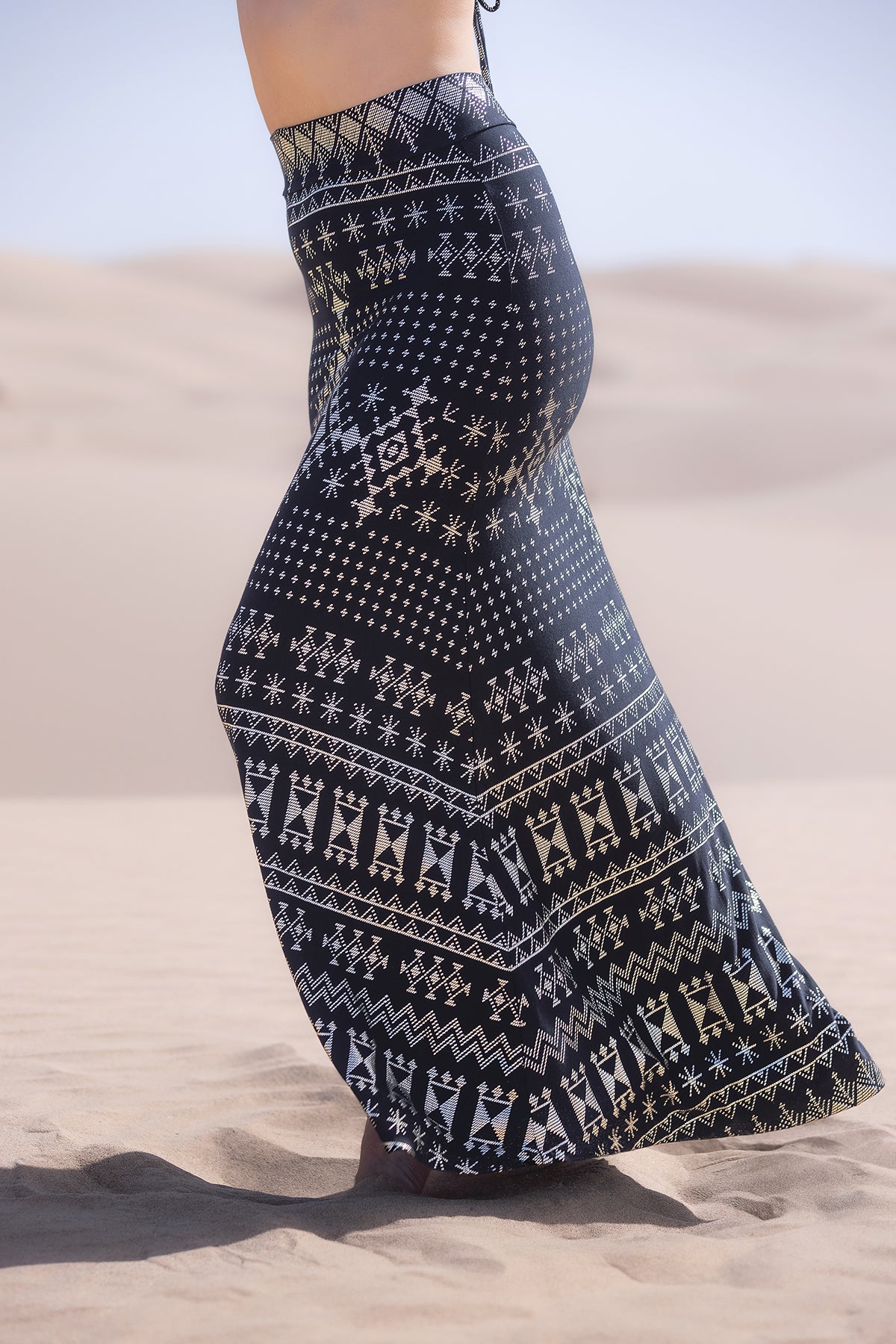 A person stands barefoot on sand wearing the Melodia Designs Alkimiya Cleopatra Maxi Skirt Obsidian, a flowing black A-line skirt with white geometric and tribal patterns, with blurred sand dunes and a clear sky in the background.
